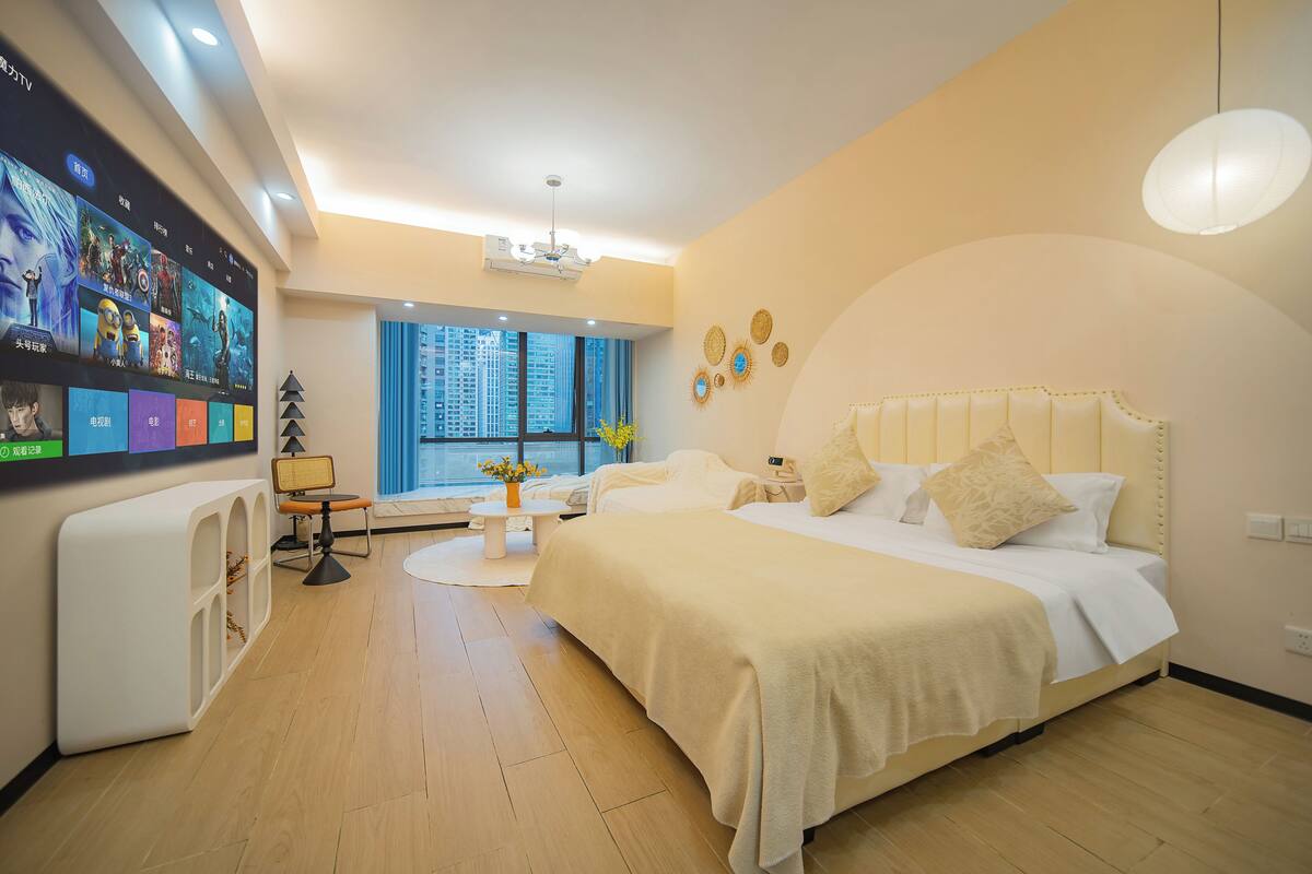 Habitación de hotel con un televisor grande que muestra carteles de películas, una cama con ropa blanca, una mesita con un jarrón de flores y vistas a edificios altos por la ventana.