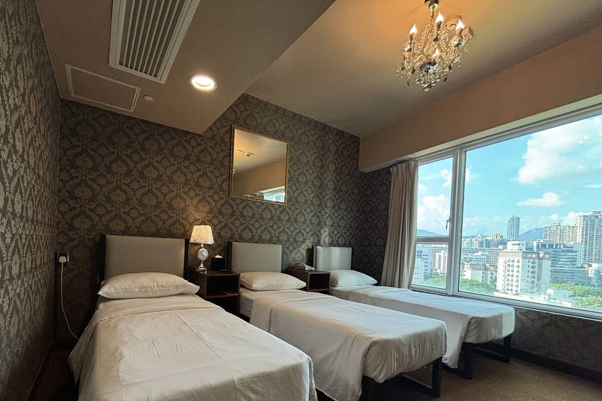 A hotel room with two beds, a large window with a city view, a chandelier, and a mirror on the wall.