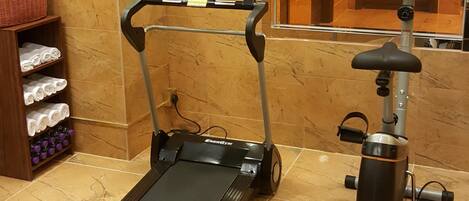 Una caminadora y una máquina elíptica en una habitación de hotel, con un espejo, un estante con toallas y un cartel decorativo de relajación.