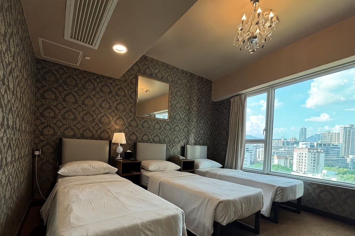 Habitació d'hotel amb tres llits, un gran finestral amb vista a la ciutat, un candelabrum i paper tapet amb dibuixos.