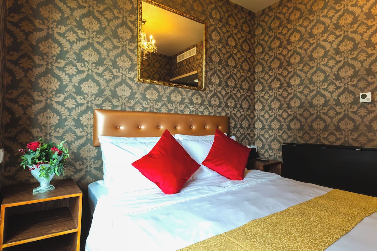 A neatly made bed with white and red pillows, a gold throw blanket, and a wooden nightstand with a vase of flowers.
