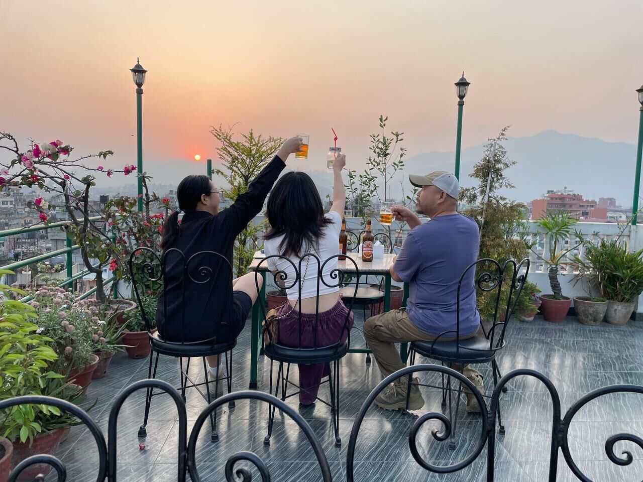 Three people seated at a table outdoors with potted plants and a sunset in the background.