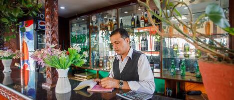Un bar con mostrador, un hombre atendiendo en la caja registradora y estantes con botellas al fondo.