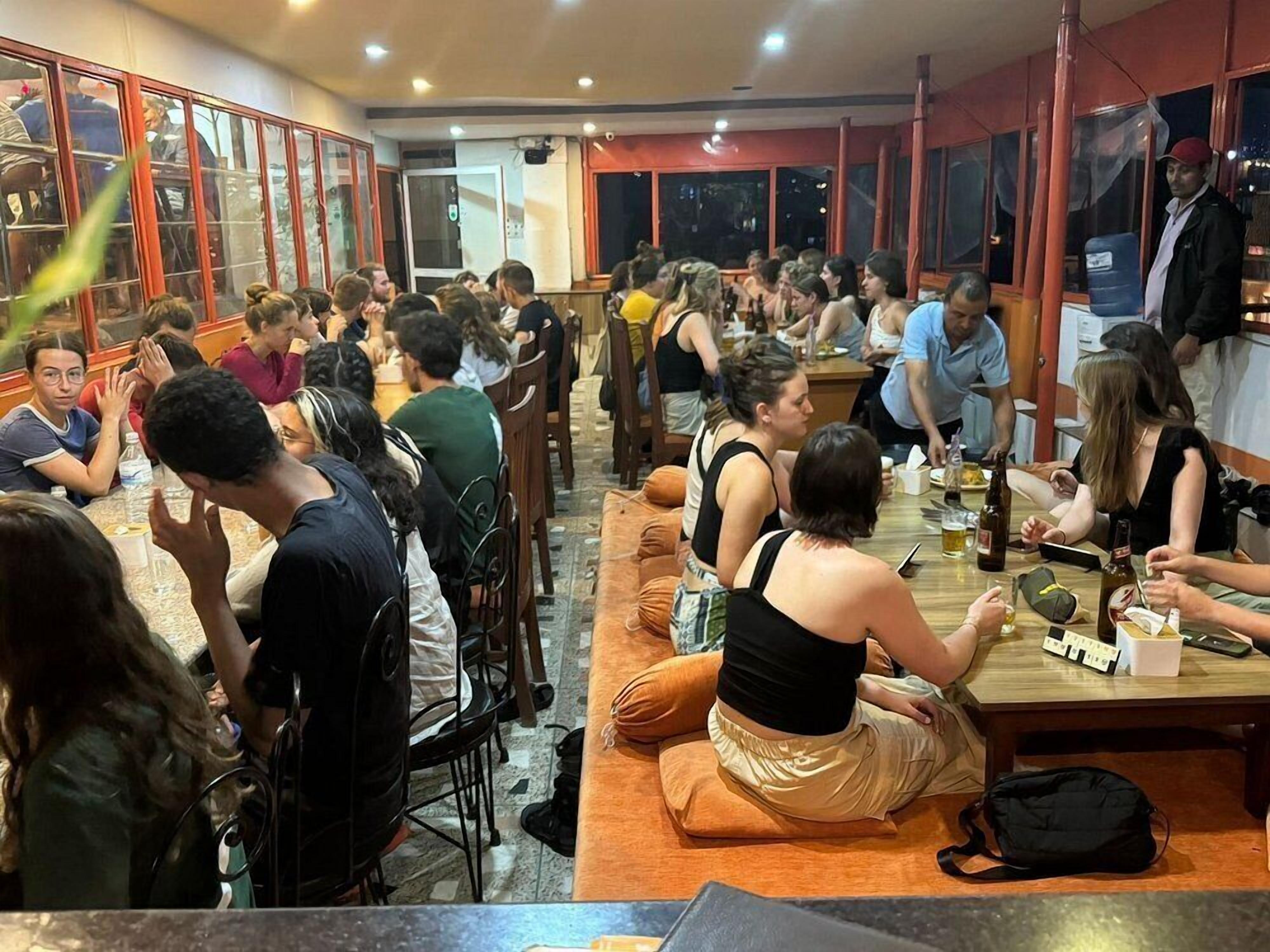 Un comedor interior concurrido con mesas y sillas, una persona de pie junto a la ventana y una puerta al fondo.