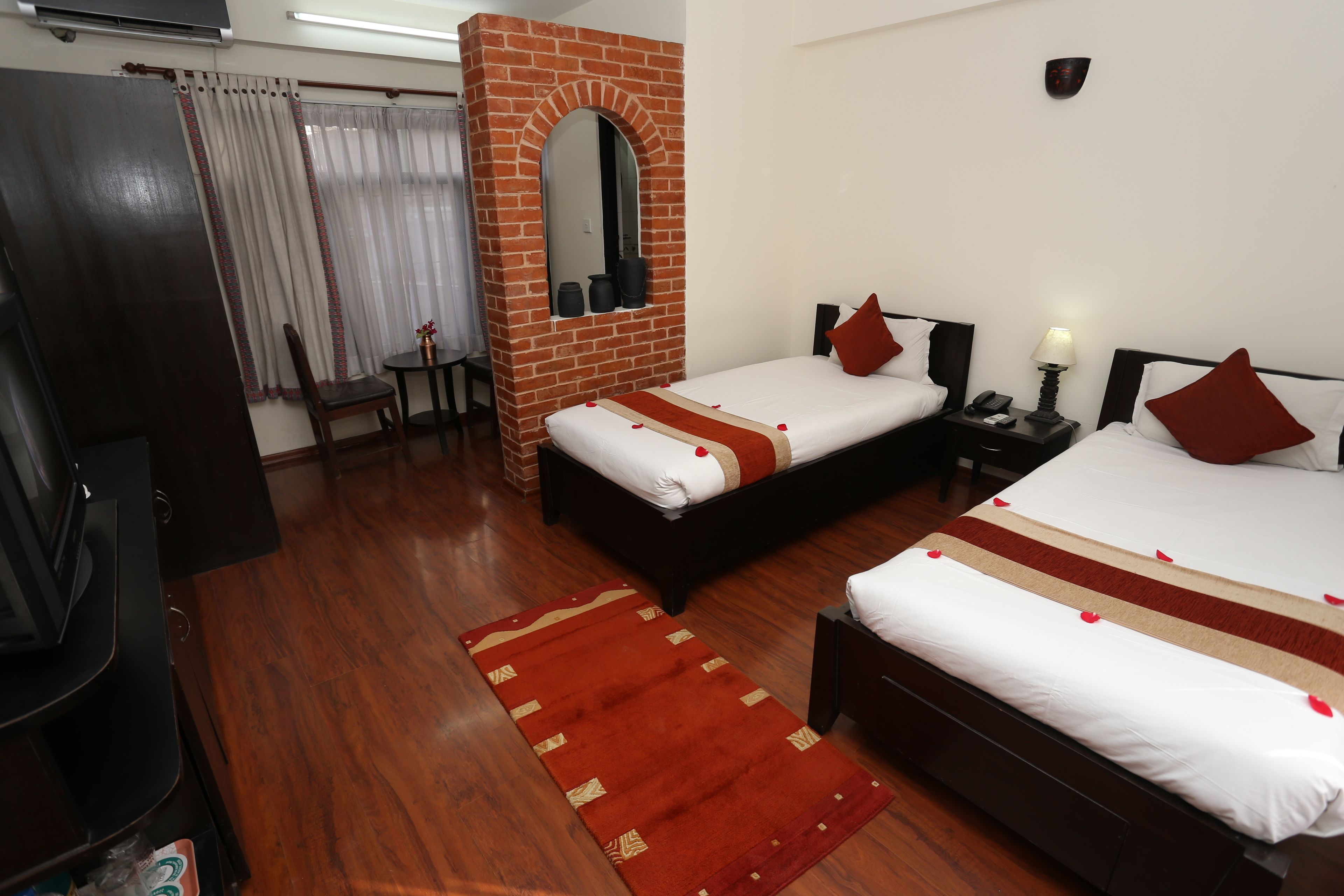 A hotel room with two beds, a wooden floor, a brick accent wall, a mirror, a small table, and a TV.