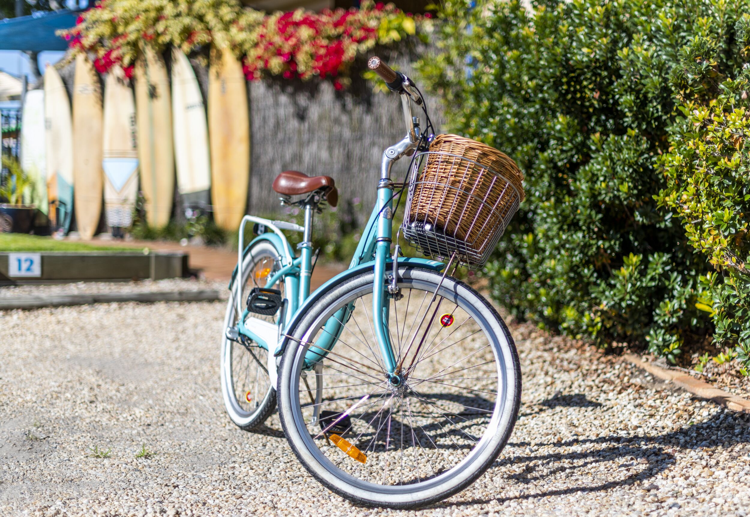 Una bicicleta color aguamarina con una cesta de mimbre, estacionada sobre un camino de grava.