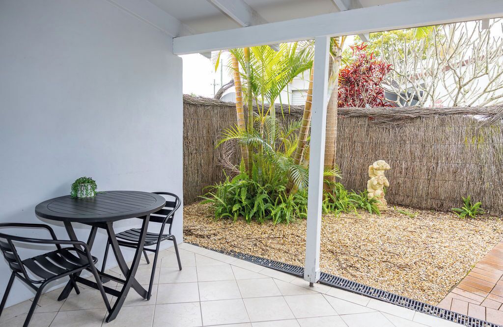 A patio with a round table and chairs, a small plant, a decorative statue, and a thatched fence.