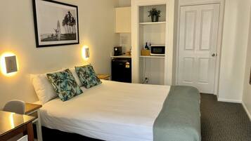 Habitación de hotel con cama, escritorio, silla, microondas, refrigerador, estante con una planta y una puerta.