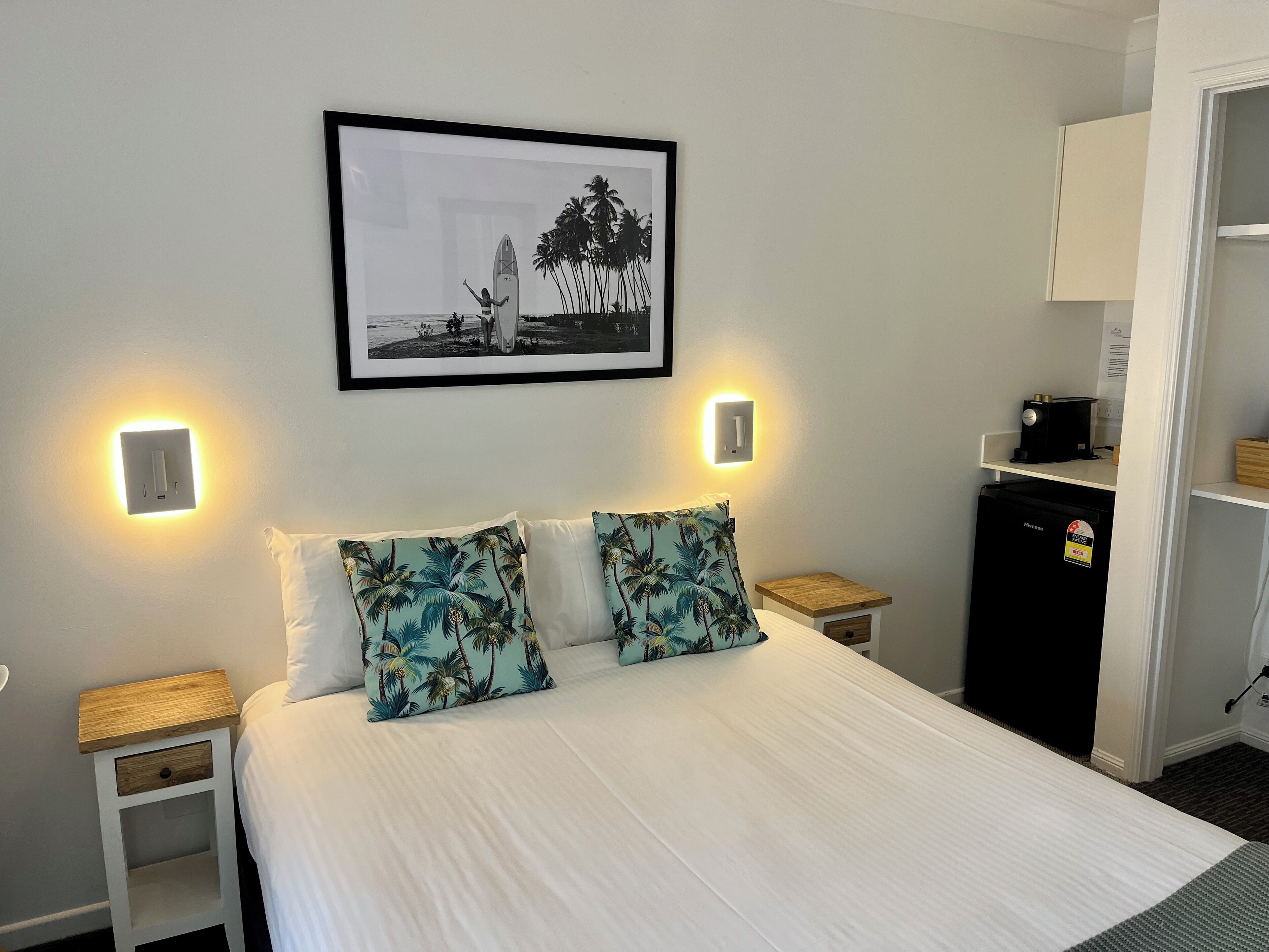 A neatly made bed with two pillows, a framed picture above, bedside tables with lamps, and a small refrigerator.