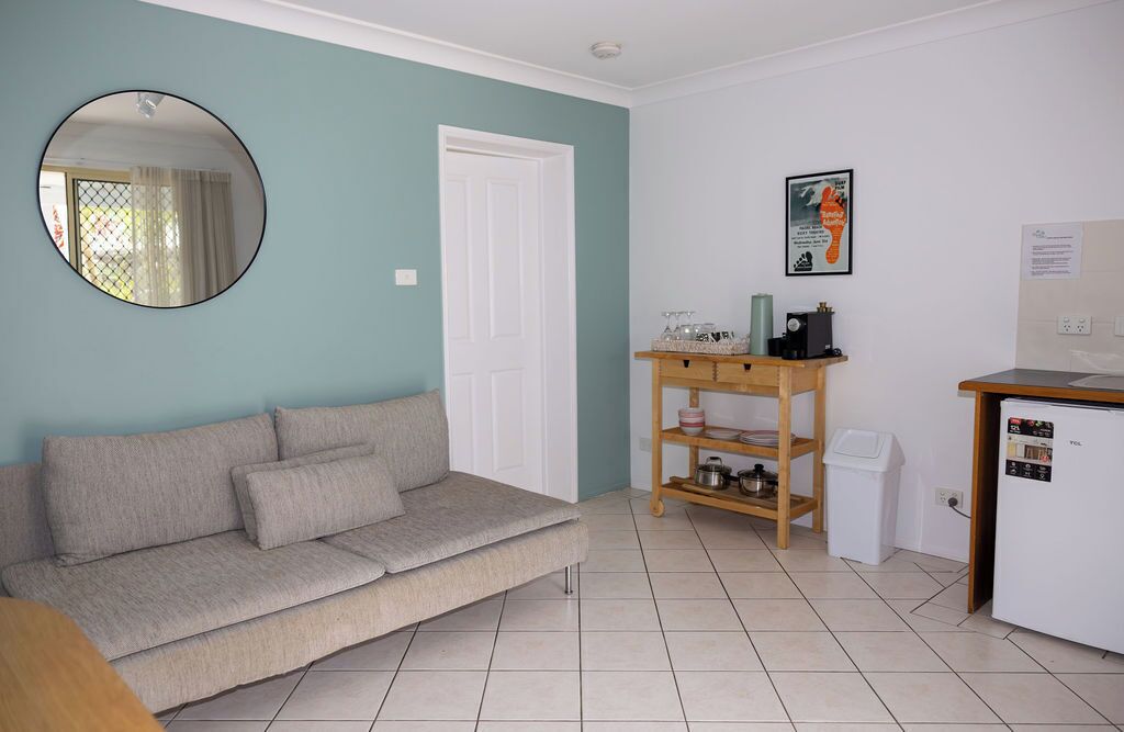 A living room with a sofa, a coffee table, a small wooden shelf with kitchen items, a white door, and a refrigerator.