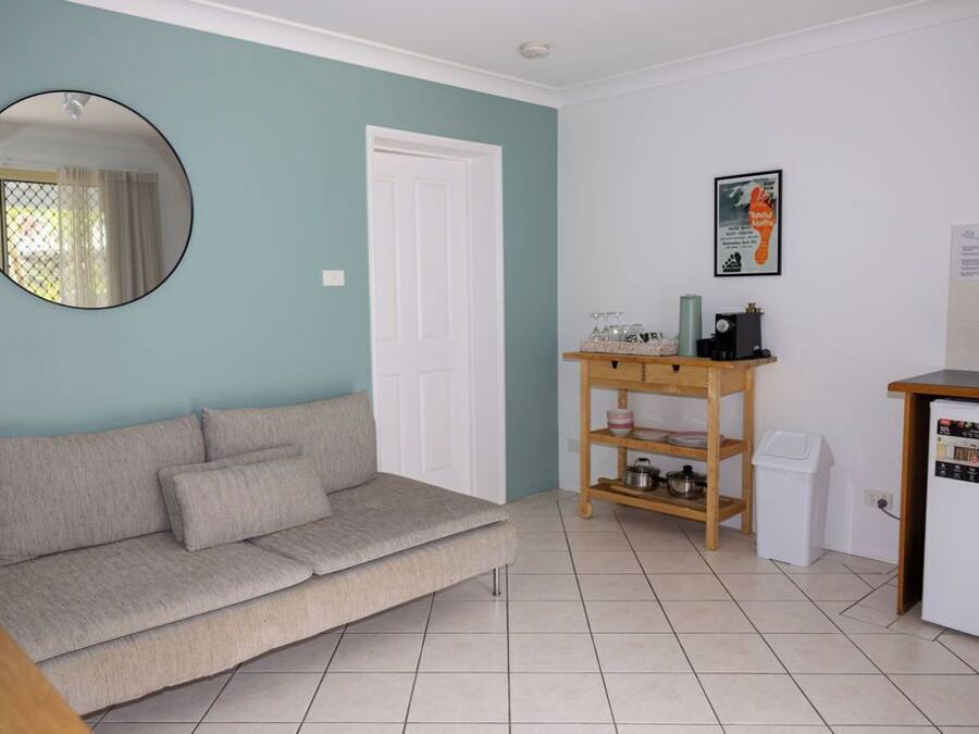 Uma sala de estar com sofá, mesa de centro, prateleira de madeira pequena com utensílios de cozinha, porta branca e geladeira.