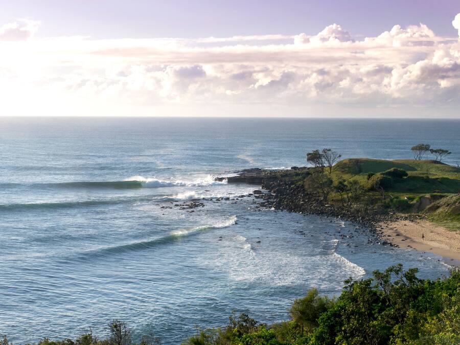 A coastal landscape with a rocky shoreline, waves, and a grassy hill with sparse trees.