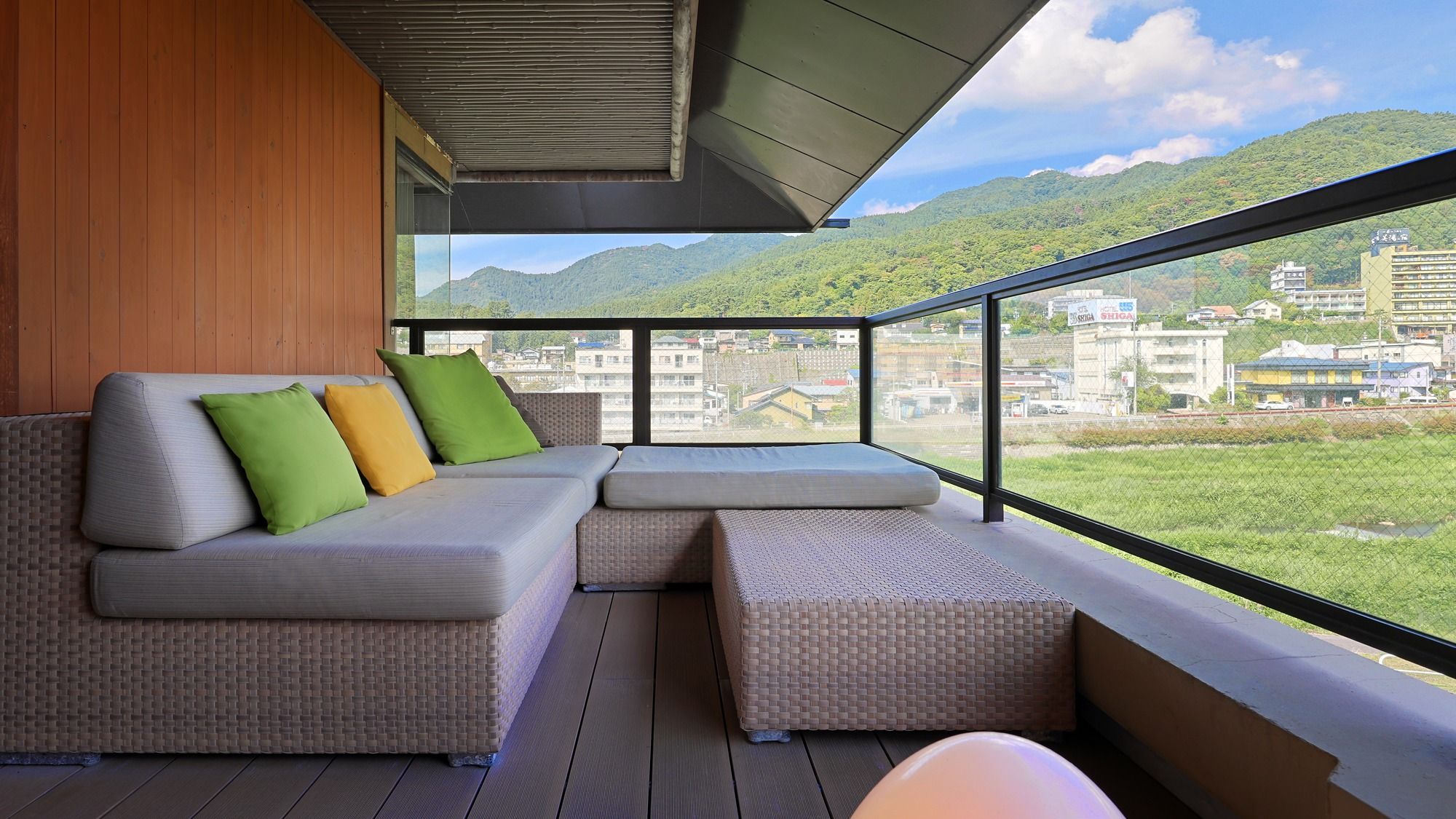 A balcony with a sofa and ottoman, a wooden wall, and a view of a cityscape and mountains.