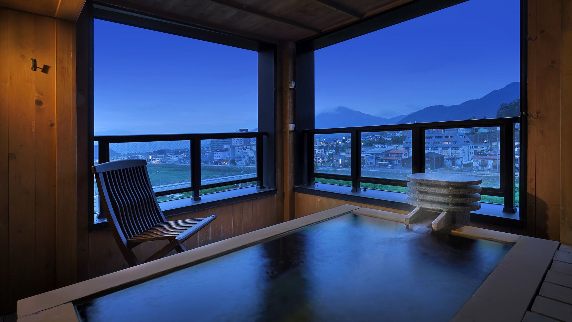Una habitación con vistas a un paisaje urbano y montañas, una silla de madera y una bañera de hidromasaje de piedra.