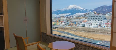 A room with tatami flooring, a low table, and chairs, featuring a large window with a view of a snow-capped mountain and a town below.