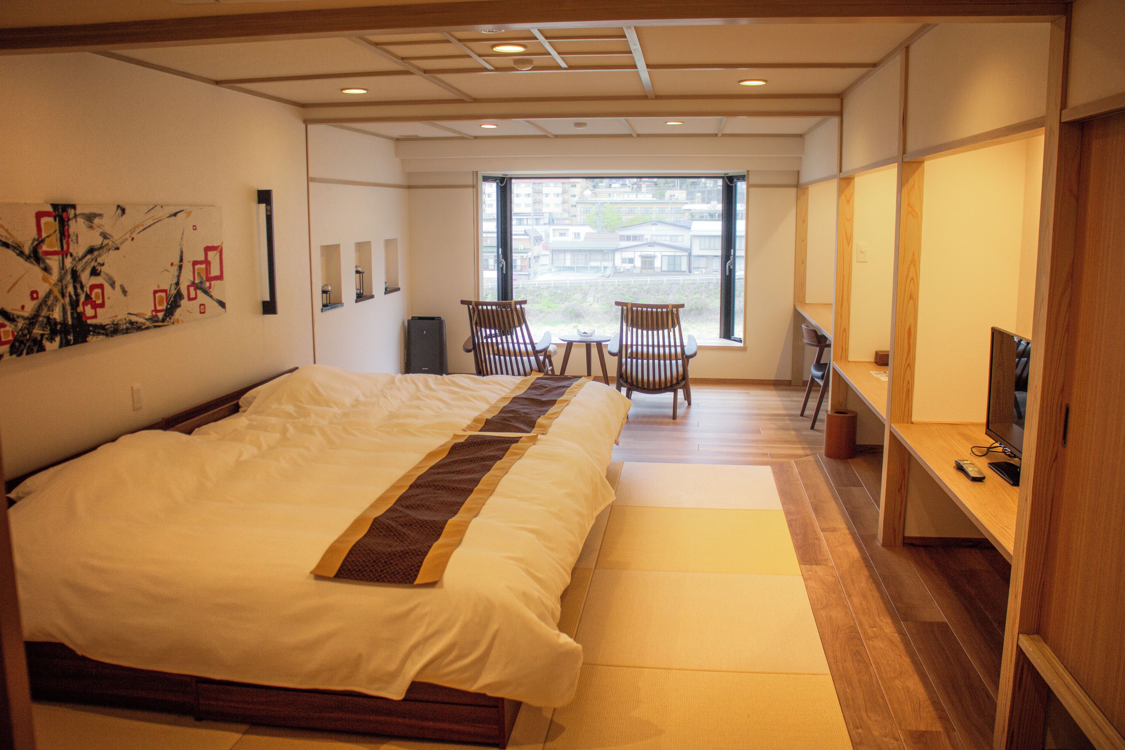 A hotel room with a large bed, a small table, two chairs, a TV, and a window with a view of buildings.