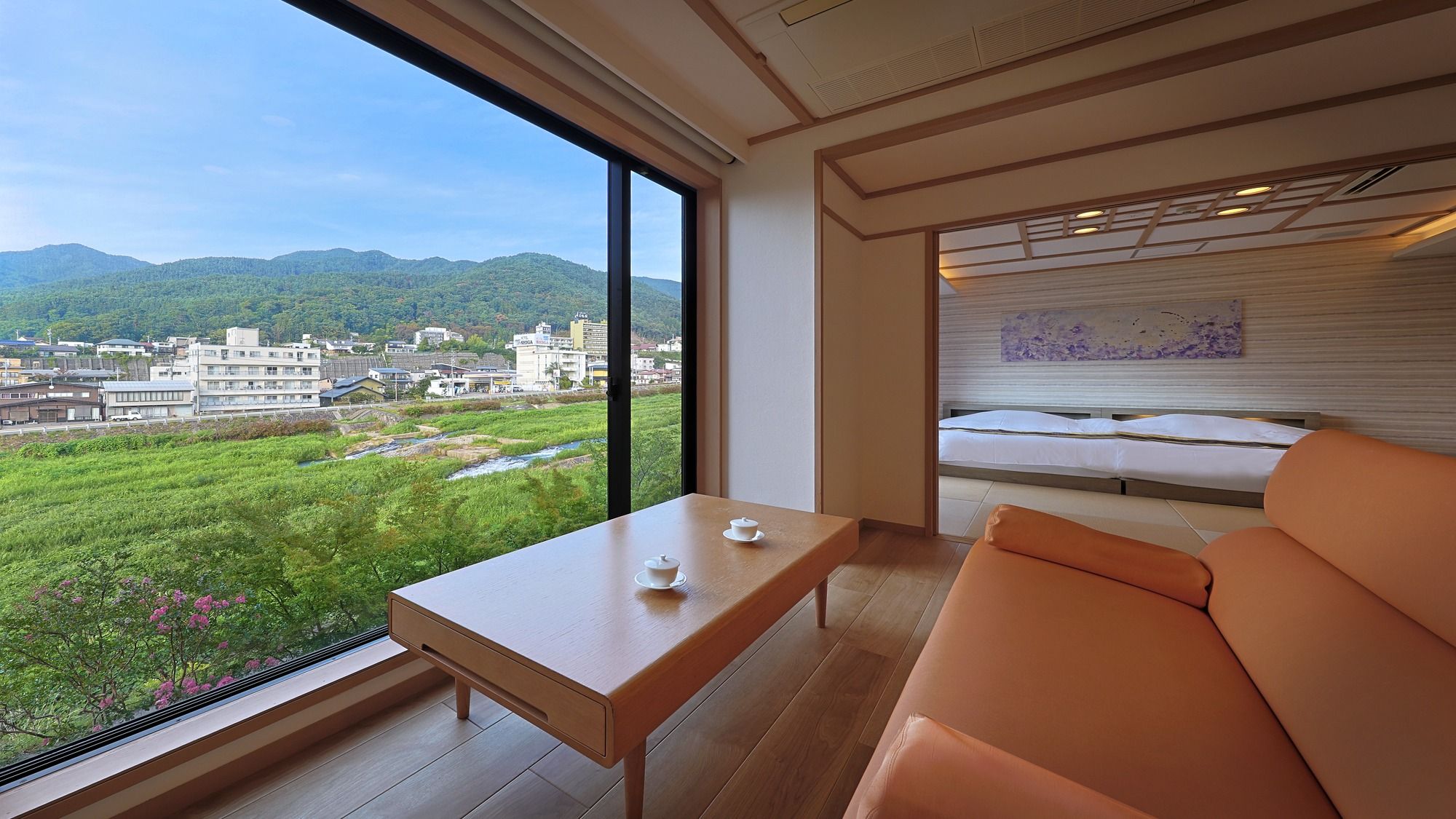 A modern room with a large window, a wooden table with two cups, a sofa, and a bed with a headboard.