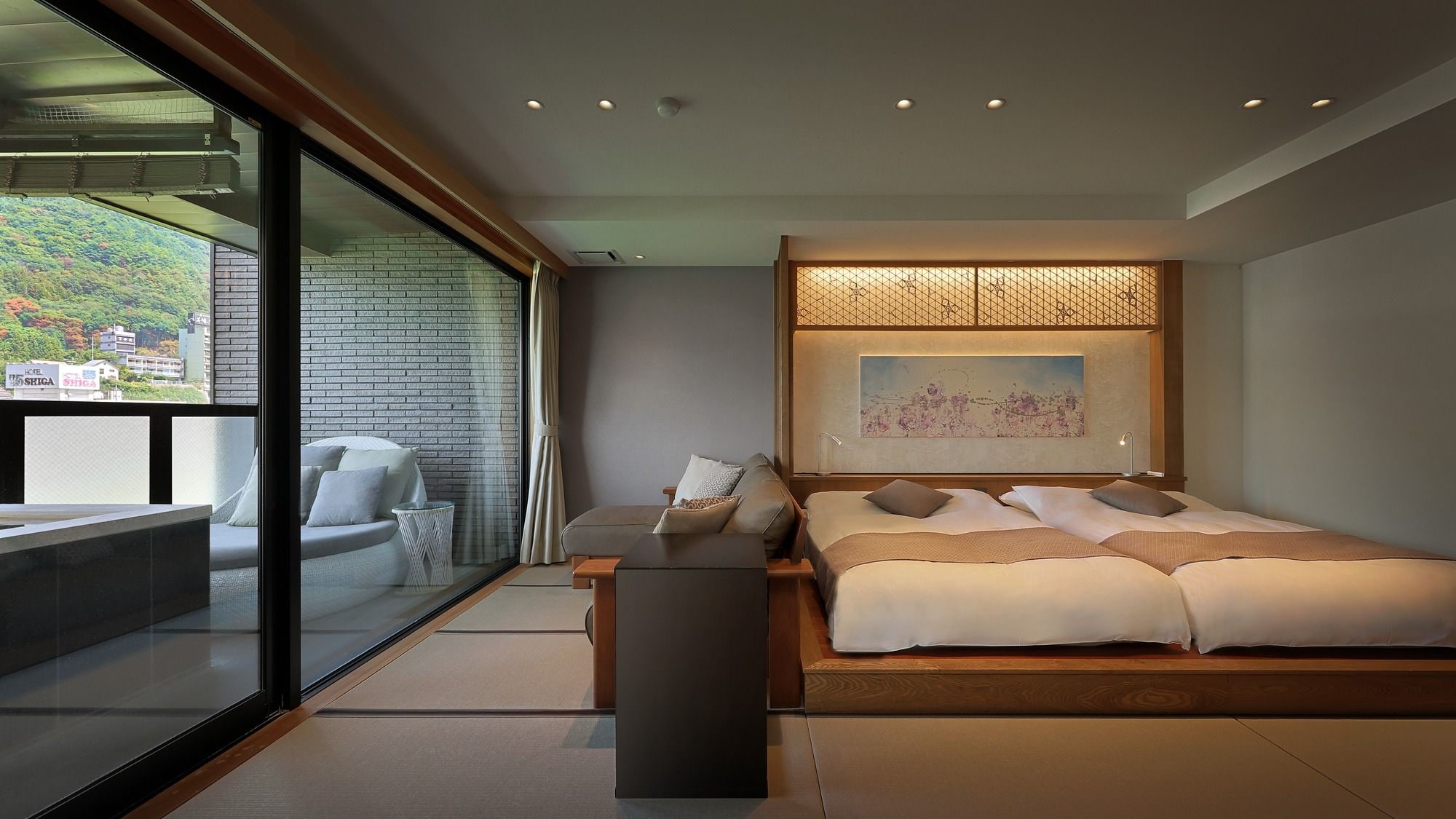 Ein modernes Schlafzimmer mit einem großen Bett, einer Sitzecke mit einer Couch und Sesseln, einer Glaswand mit Blick auf einen grünen Hügel und einer Holzbalkendecke.
