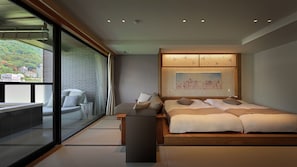 A modern bedroom with a large bed, a seating area with a sofa and armchairs, a glass wall with a view of a green hillside, and a wooden ceiling.