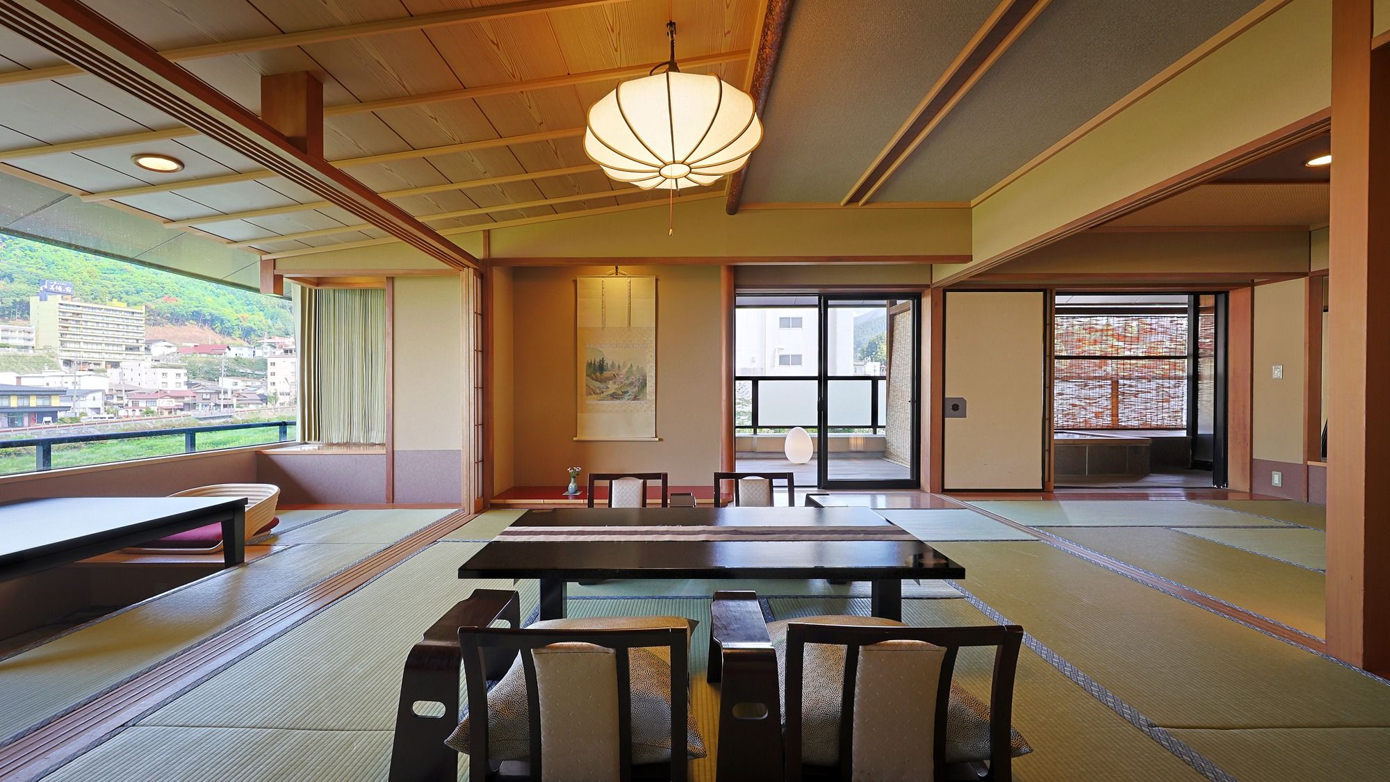 A traditional Japanese room with tatami flooring, a low table, chairs, a hanging light fixture, and a view of a cityscape through a large window.
