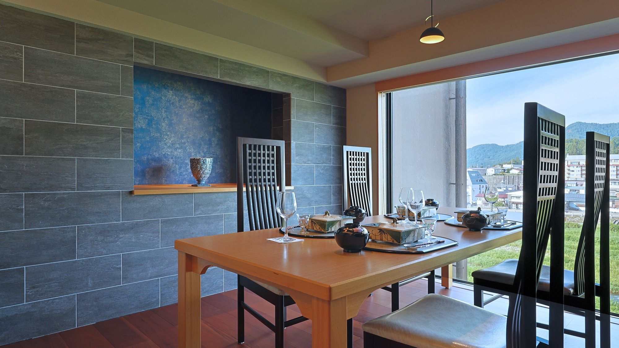 Un comedor con una mesa lista para dos, vista a la ciudad a través de amplios ventanales, y una decoración moderna con paredes de piedra oscura y una mesa de madera.