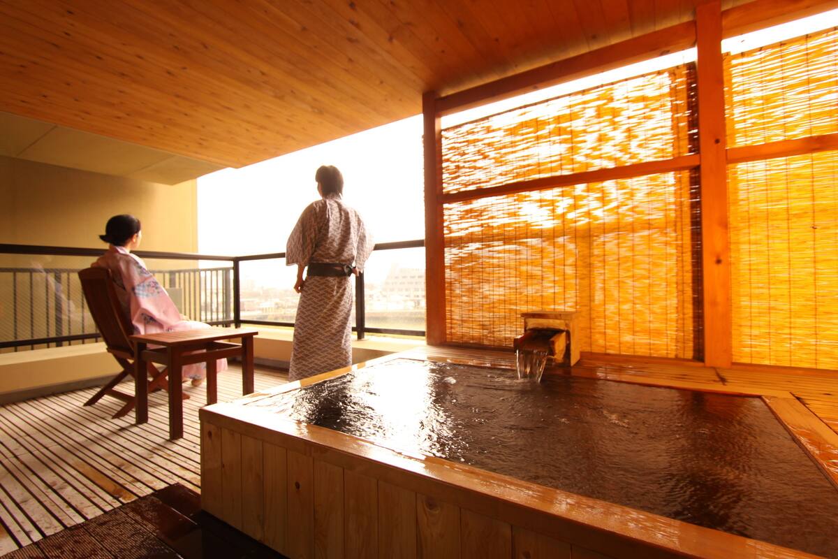 Un onsen japonais traditionnel, avec un intérieur en bois, un bain d’eau chaude et une vue dégagée sur l’extérieur grâce à une grande fenêtre.