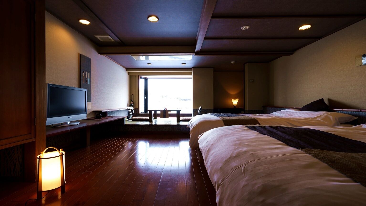 A hotel room with two beds, a TV, a desk, a lamp, and a view of the outdoors.