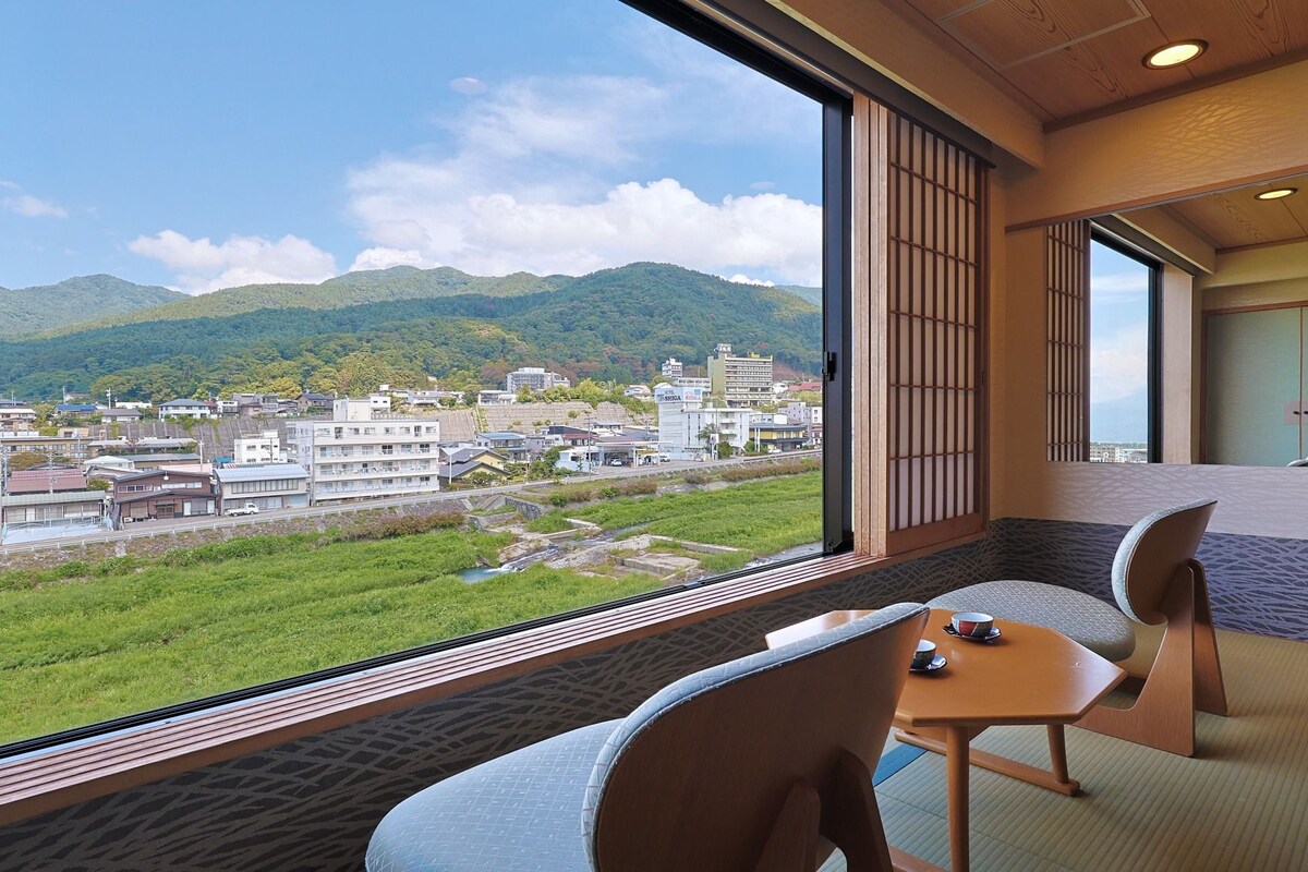A room with a view of a cityscape and mountains, featuring a traditional Japanese sliding door, tatami flooring, and a low table with a cup.