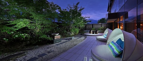 An outdoor seating area with a sofa and chairs, surrounded by greenery and a gravel path.