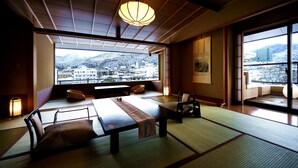 Una habitación japonesa tradicional con suelo de tatami, una mesa baja y cojines. Un ventanal ofrece vistas a un paisaje urbano y montañas.