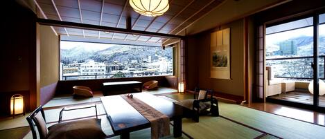 Una habitación japonesa tradicional con suelo de tatami, una mesa baja y cojines. Un ventanal ofrece vistas a un paisaje urbano y montañas.