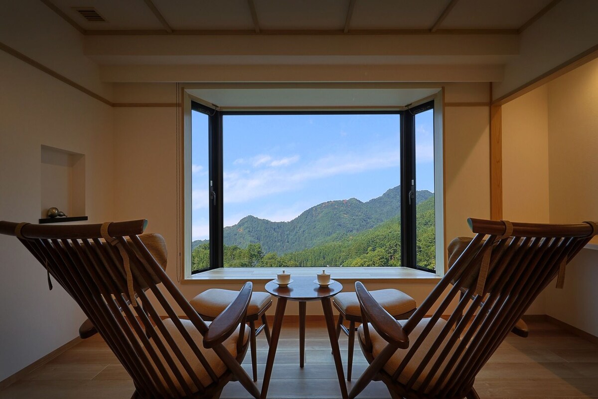 A room with a large window, two wooden chairs, a small table, and a view of a mountain.
