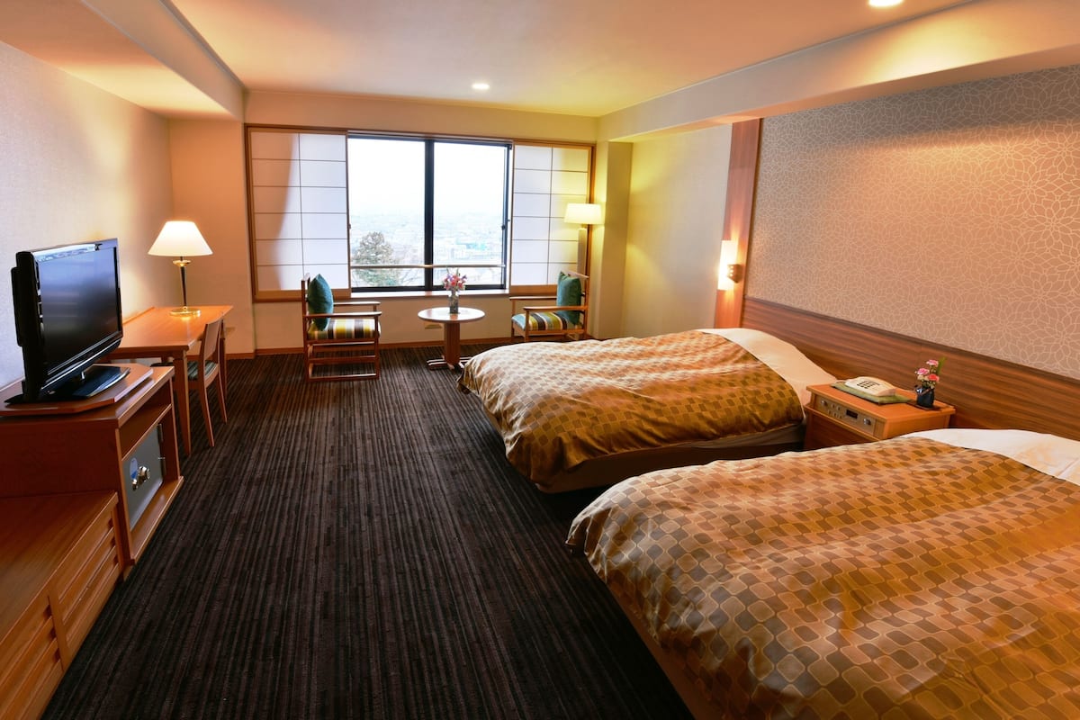 A hotel room with two beds, a desk with a television, a small table with a lamp, and a window with a view.