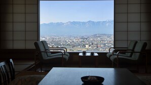 Una habitación con un ventanal amplio que ofrece vistas a una ciudad y montañas. Hay dos sillones y una mesa con un cuenco encima.