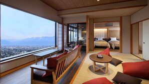 Una habitación de hotel moderna con un amplio ventanal que ofrece vistas a la ciudad, una zona de estar con un banco de madera y cojines, una pequeña mesa redonda, y un dormitorio separado con una cama y una mesita de noche.