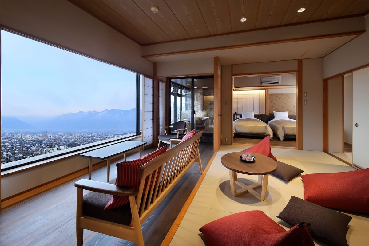 A modern hotel room with a large window offering a city view, a seating area with a wooden bench and cushions, a small round table, and a separate bedroom with a bed and a nightstand.