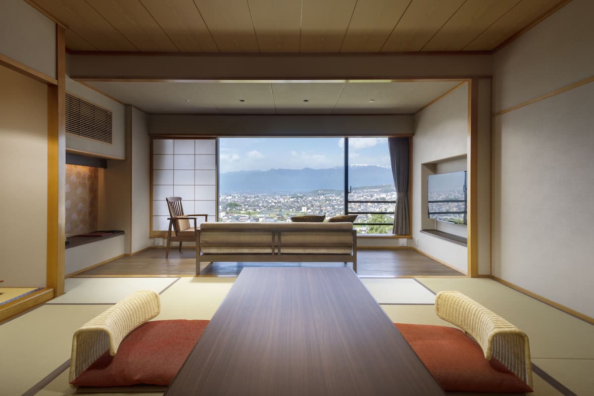 A traditional Japanese room with tatami flooring, a low table, two cushions, a chair, and a view of a cityscape through large windows.