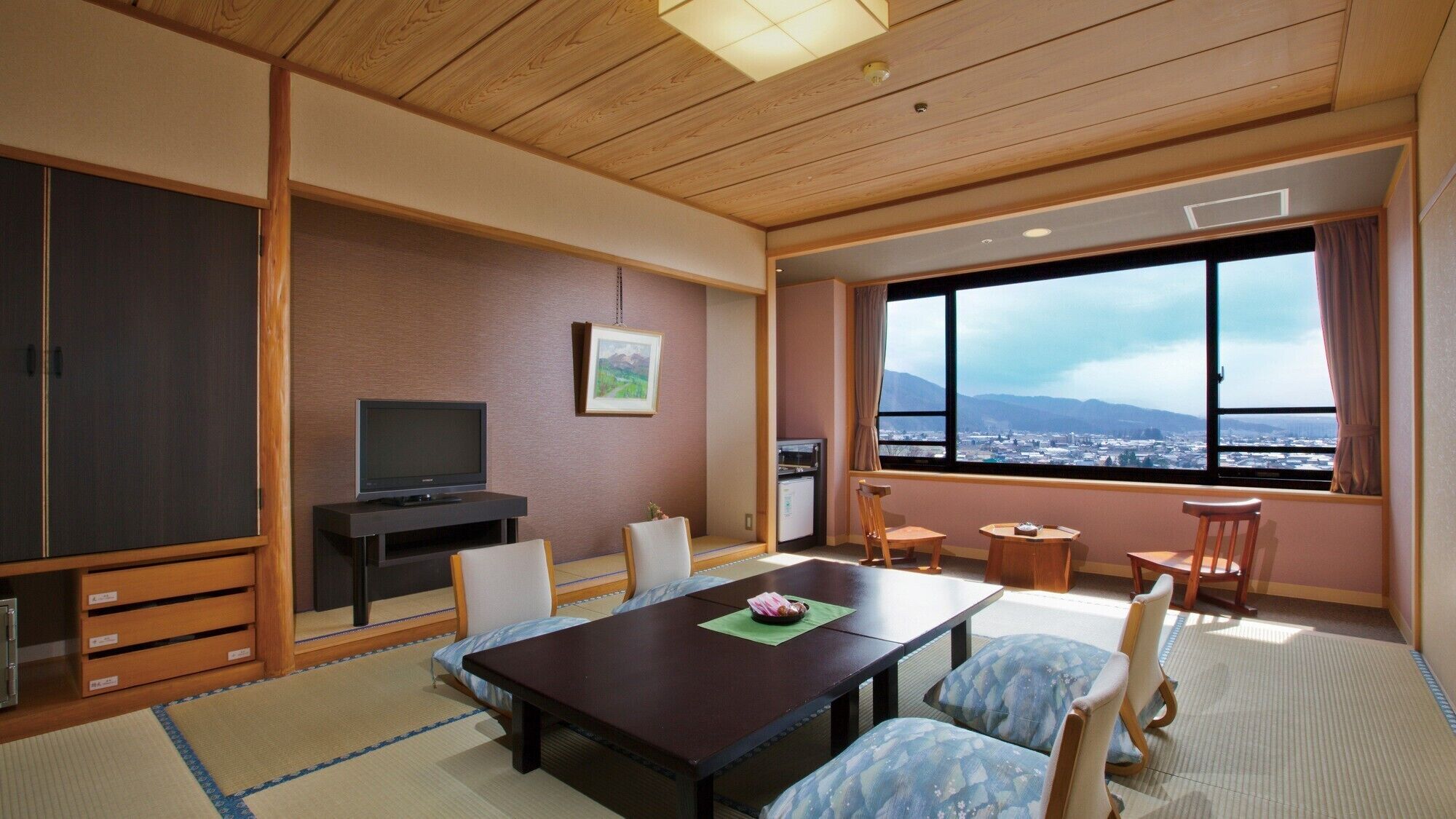 A hotel room with a wooden ceiling, a television, a dining table with chairs, a small kitchen area, and a large window with a city view.