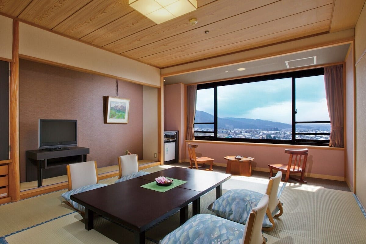 A hotel room with a wooden ceiling, a television, a dining table with chairs, a small kitchen area, and a large window with a city view.