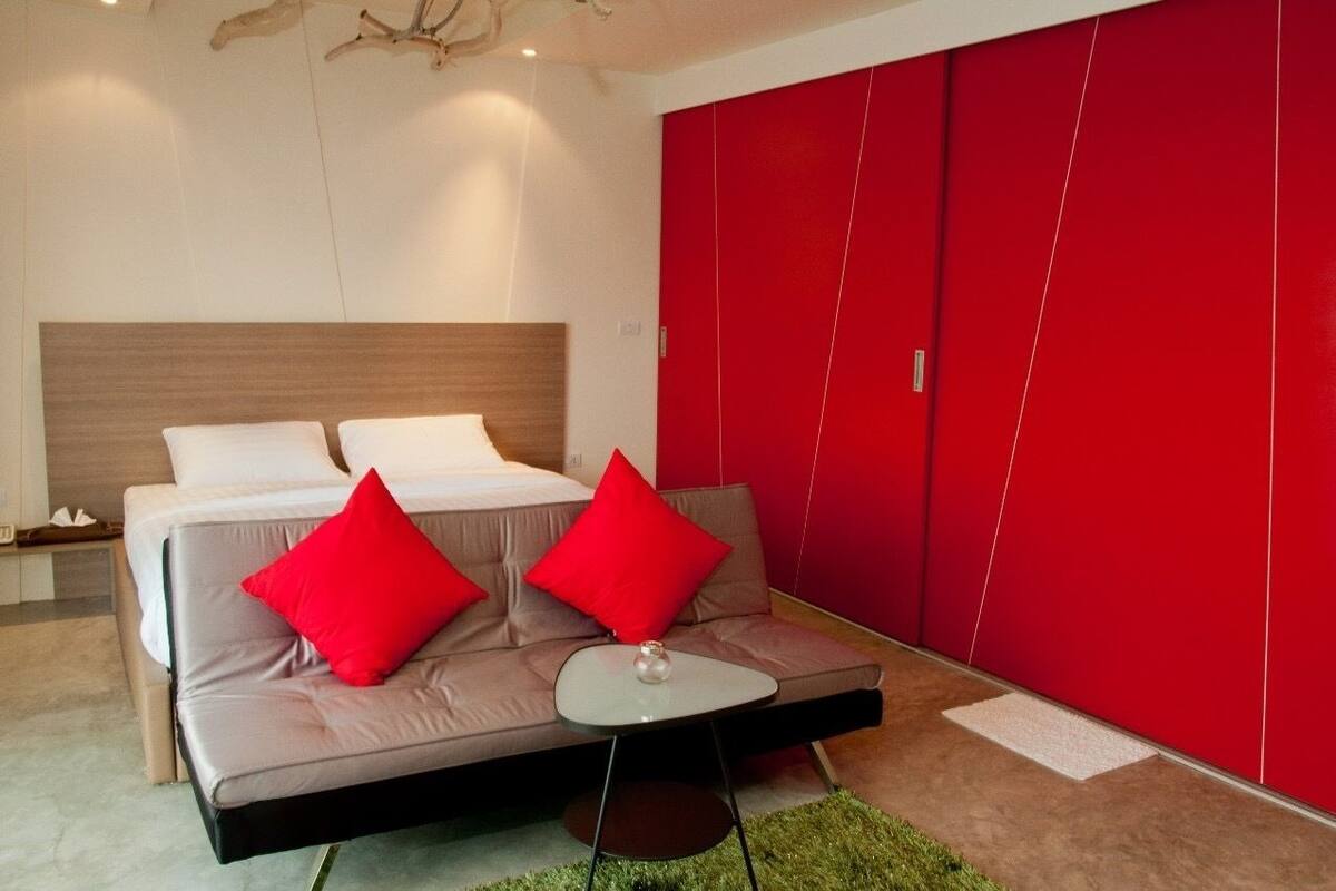 Ein modernes Schlafzimmer mit einem Bett, einem grauen Sofa mit roten Kissen, einem kleinen Tisch und einem roten Kleiderschrank.