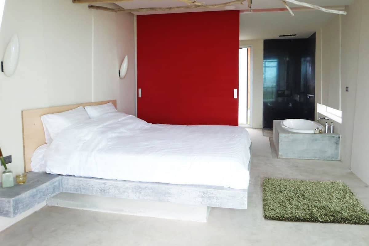 Ein modernes Schlafzimmer mit einem großen Bett, einer roten Tür, einem Badezimmer mit Waschbecken und Spiegel sowie einem einzigartigen Deckengestalt.