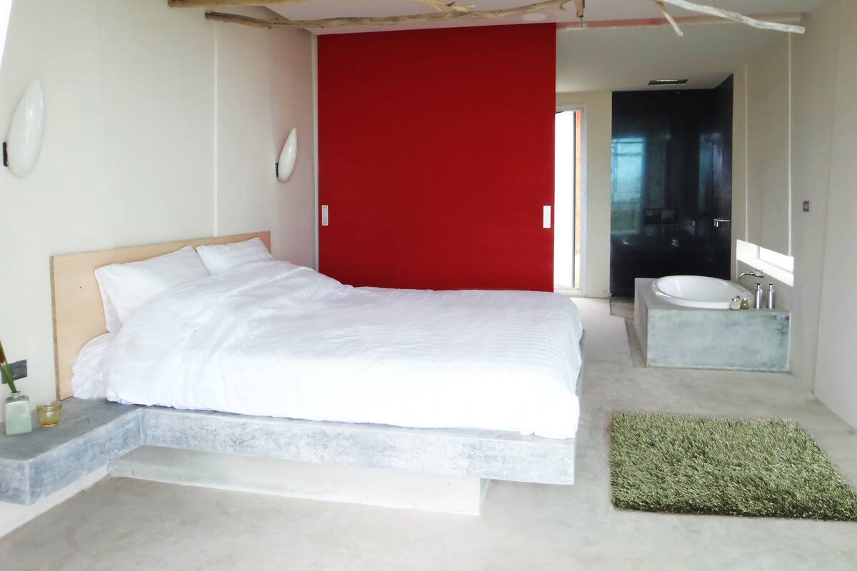 Ein modernes Schlafzimmer mit einem großen Bett, einer roten Akzentwand, einem Badezimmer mit Waschbecken aus Marmor und einem einzigartigen Deckendesign.