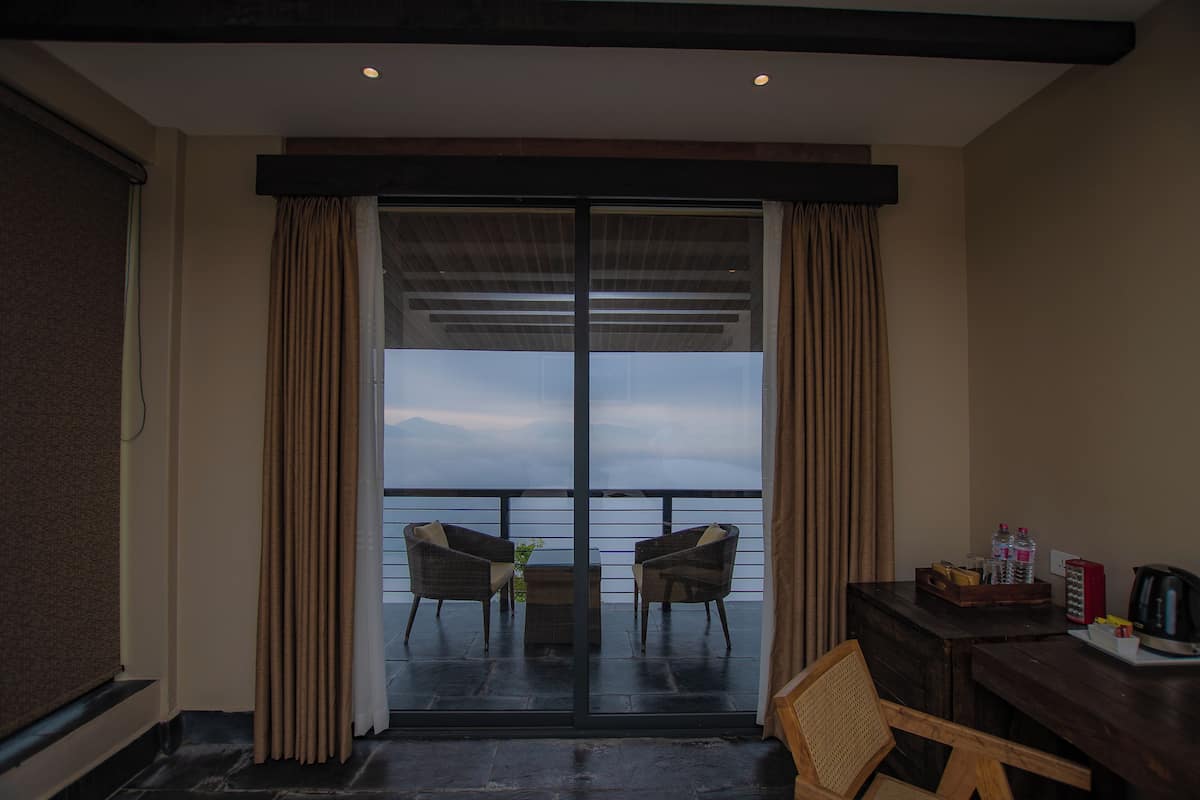 A room with a balcony, two wicker chairs, a small table, a wooden chair, and a desk with a kettle and water bottles.
