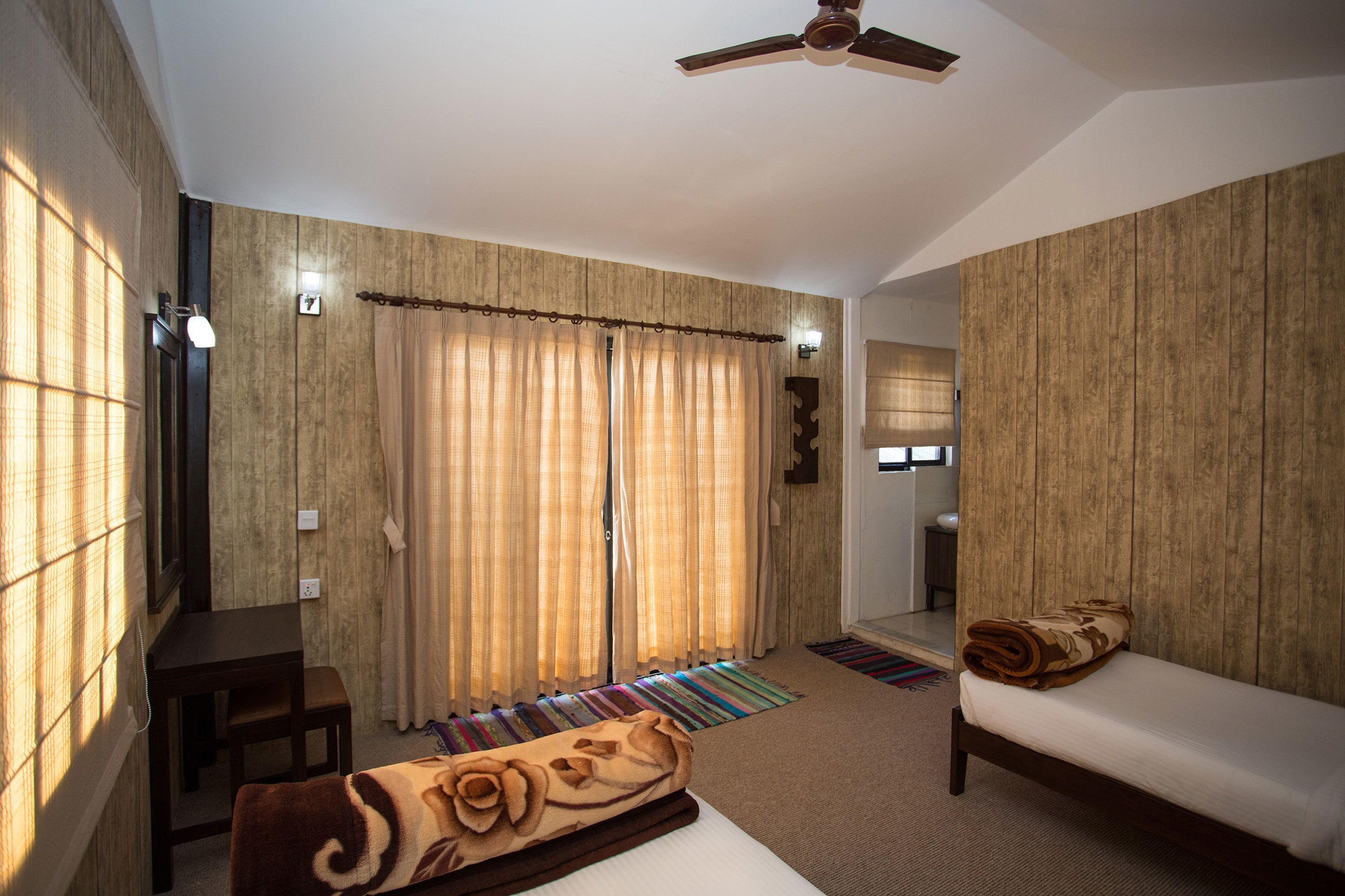 A room with a bed, a bench, a small table, a ceiling fan, and curtains.