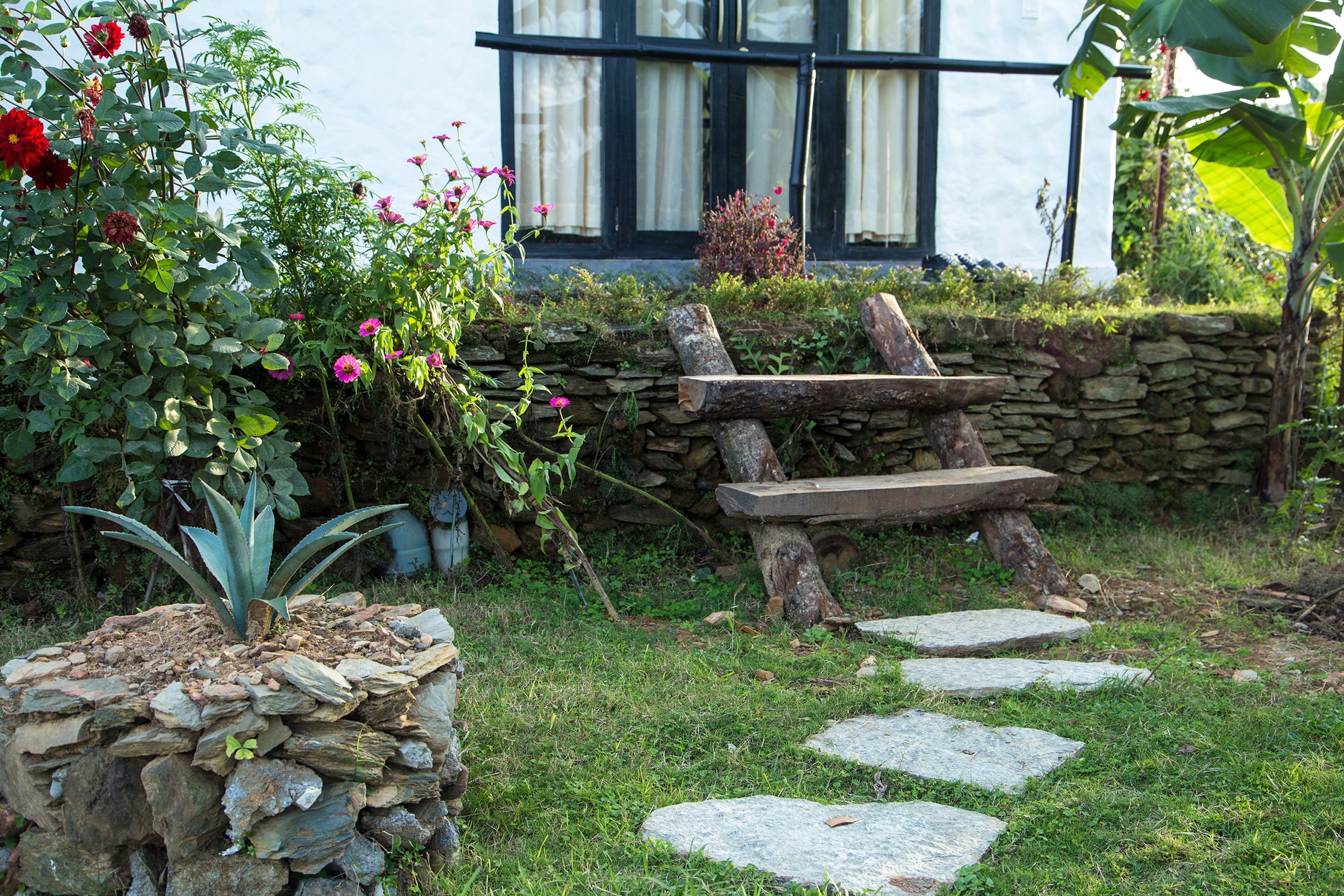 A stone bench in a garden with a stone wall and steps leading to a building with windows.