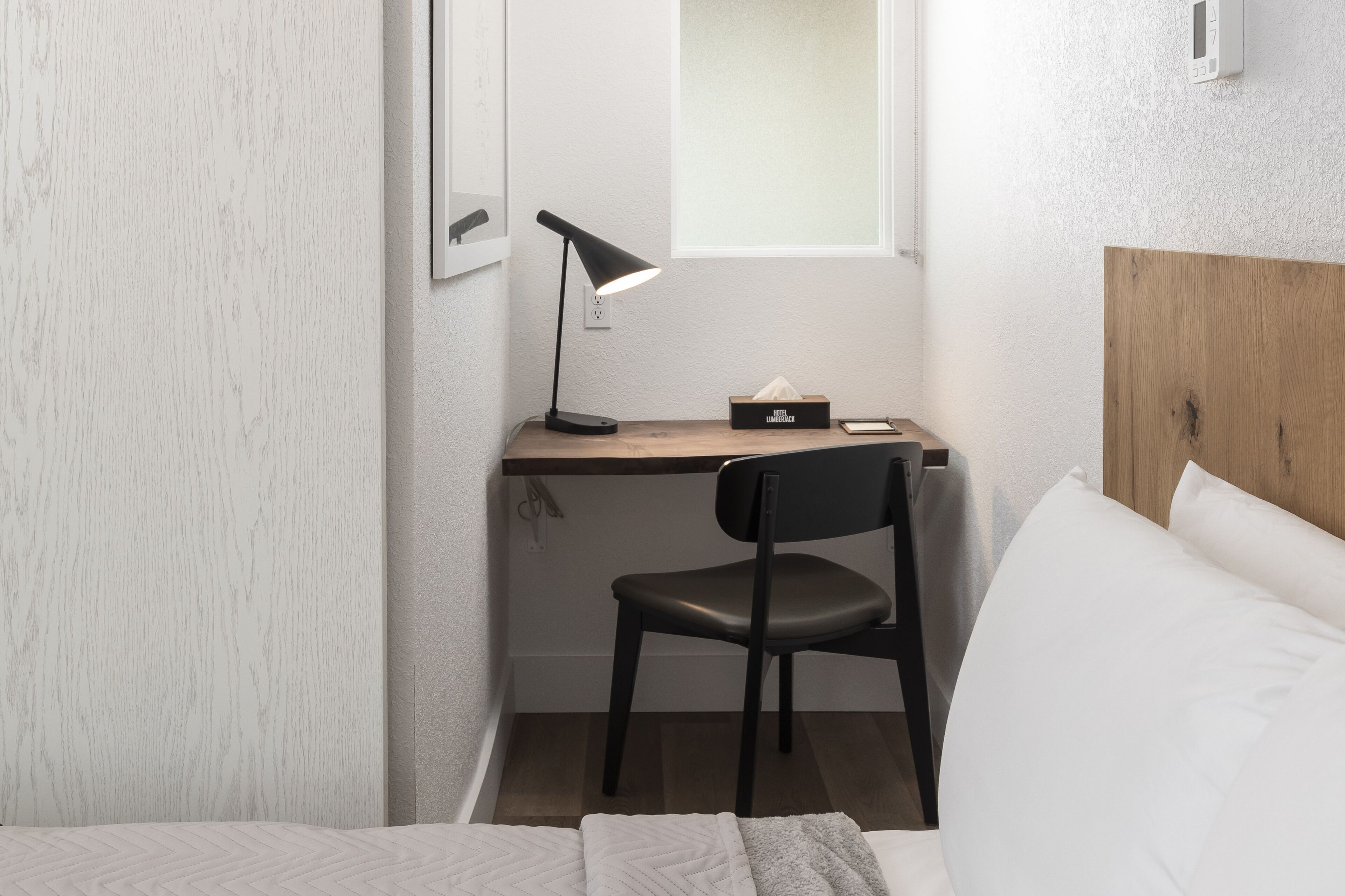 A small, neatly arranged bedroom with a wooden desk, a chair, a bedside table, and a bed.