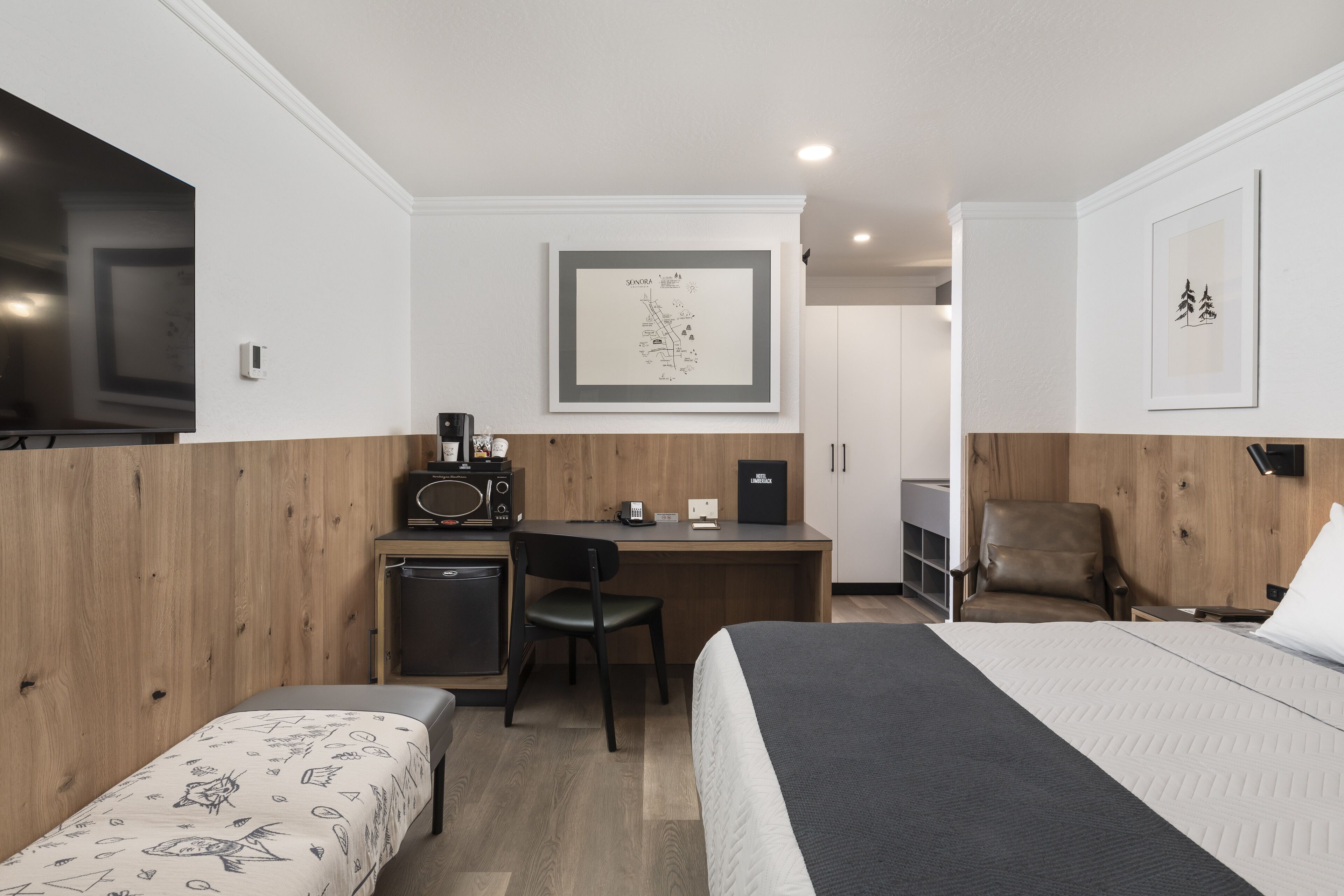 A hotel room with a bed, a desk, a chair, a small table, a TV, and a framed map on the wall.