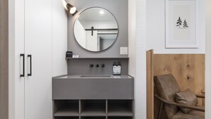 Un baño con tocador gris, un espejo redondo, una silla de madera y una obra enmarcada en la pared.