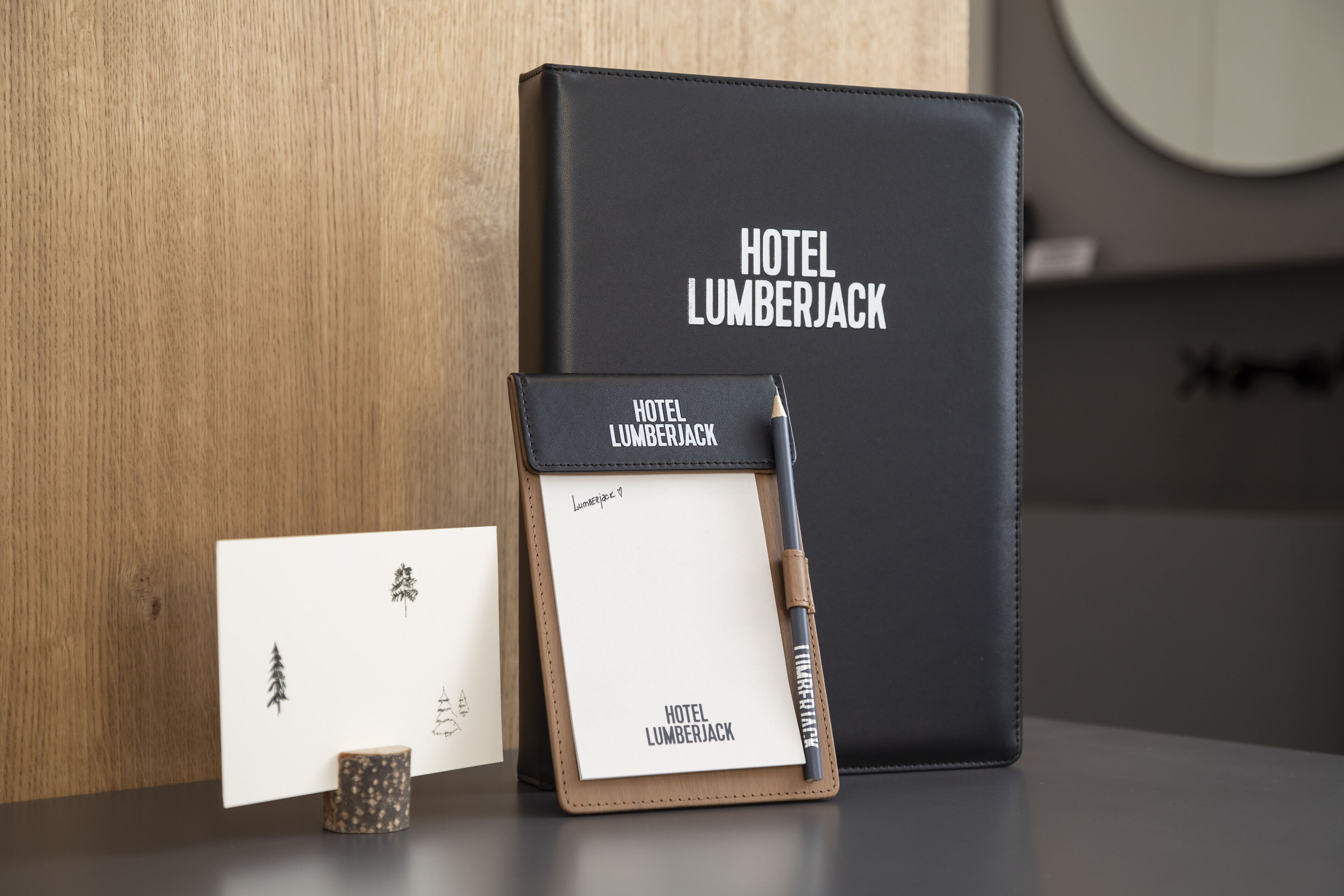 A black leather-bound notebook with 'HOTEL LUMBERJACK' embossed on the cover, a pencil, and a small wooden coaster with a tree design.