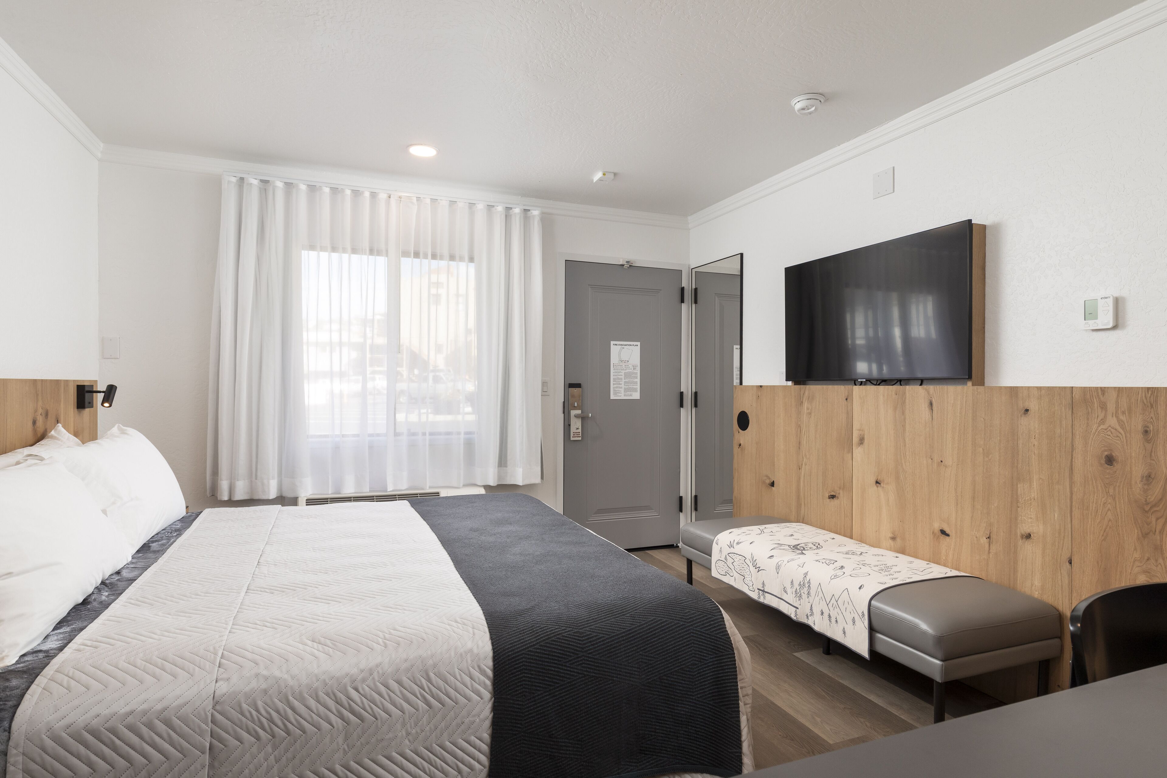 A hotel room with a bed, a bench, a TV mounted on the wall, and a door.
